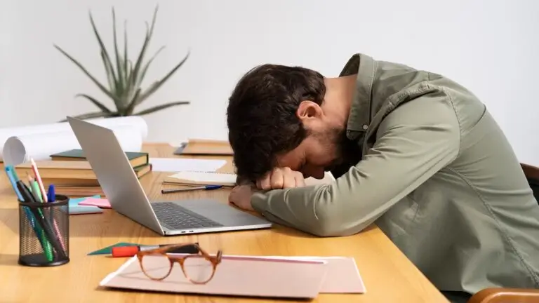 Hombre cansado y estresado, durmiendo sobre un escritorio de trabajo con portátil, documentos y material de oficina, reflejando agotamiento laboral y burnout.